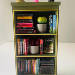 May include: A miniature green bookshelf with yellow trim, filled with small books, potted plants, and decorative figurines. The books include titles like "Bravely", "Stranger Things", and "Goosebumps". A small desk lamp sits on top.