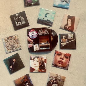 May include: A collection of miniature vinyl record albums and a Zuru Mini Brands box featuring the text Vinyl Vinyle. The set includes various square album covers with artists such as Kiss, Shania Twain, Sting, and The Mamas and the Papas.