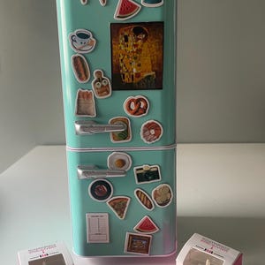 May include: A miniature teal refrigerator with various food-themed magnets, including ice cream cones, a watermelon slice, and a painting. Three small cakes and two miniature roasted chickens are in front of the refrigerator.
