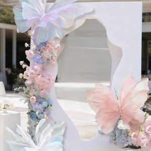 May include: A white, abstract frame decorated with large, translucent butterfly wings in pastel colors. The frame is adorned with pink, blue, and white flowers. The butterflies have pearl accents, creating a whimsical and elegant display.