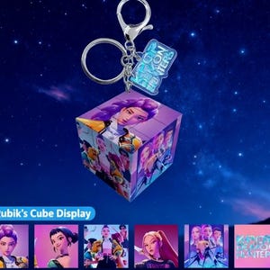 May include: A Rubik's Cube display keychain featuring a purple-haired woman on multiple sides. The keychain has a silver clasp and a small, rectangular tag with the text "KPOP DEMON HUNTERS". The cube is surrounded by a dark blue background with stars.