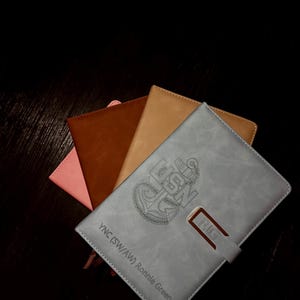 May include: A collection of leather-bound notebooks in various colors: pink, brown, tan, and gray. The gray notebook features an engraved US Navy emblem, the text "YNC (SW/AW) Ronnie Green", and a metal clasp with the letters "RIG".