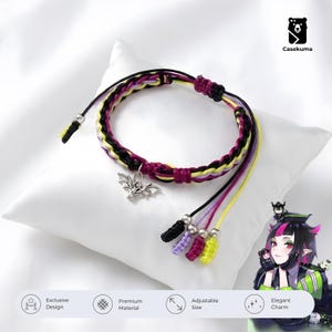 May include: A braided bracelet with black, purple, yellow, and pink threads. It features a silver bat charm and dangling threads with decorative beads. The bracelet is displayed on a white pillow.