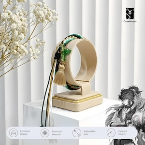 May include: A beige bracelet display stand with a gold trim, holding a colourful bracelet. The bracelet features green, black, and white accents, a green charm, and a tassel. The stand is on a white surface.