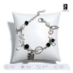 Könnte beinhalten: Silberfarbenes Charm-Armband mit schwarzen und weißen Perlen, klaren Kristallakzenten sowie Stern- und Flügel-Anhängern. Ein kleiner Buch-Anhänger ist ebenfalls enthalten. Das Armband wird auf einem weißen Kissen präsentiert.