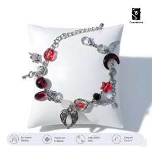 Op de afbeelding: Zilverkleurige armband met diverse bedels, waaronder vleugels, een sleutel, sterren en kralen in rode, zwarte en transparante kleuren. De armband wordt gepresenteerd op een wit kussen. De armband heeft een verstelbare maat.