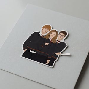Op de afbeelding: Een sticker met drie personages uit de Harry Potter-serie. De personages zijn afgebeeld in hun Hogwarts-uniformen, met zwarte gewaden, rode stropdassen en een toverstaf. De sticker staat op een grijze achtergrond.