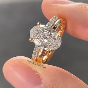 Peut inclure: Une bague en or avec une grande pierre centrale taille ovale entourée de petites pierres serties pavé sur l'anneau. La bague présente un design à tige fendue avec une finition en or poli. Le texte RIVAAJEWELSIN est imprimé sur le côté de l'anneau.