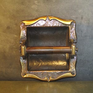 May include: Ornate, antique-style toilet paper holder with a bronze finish. The holder features a rectangular frame with decorative floral and leaf patterns, and a gold-colored trim. The roller is a dark brown color.