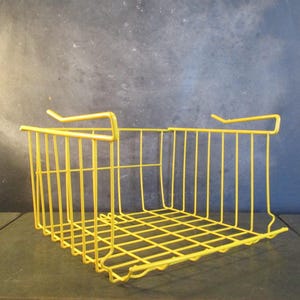 Könnte beinhalten: Ein leuchtend gelber Drahtkorb mit rechteckiger Form. Der Korb hat einen Gitterboden und vertikale Stäbe, die die Seiten bilden. Er verfügt über zwei gebogene Haken zum Aufhängen. Das Design ist schlicht und funktional.