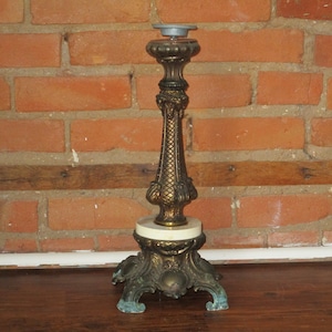 Puede incluir: Un candelero de metal dorado con una base de mármol blanco y detalles ornamentados. El candelero tiene una parte superior redonda y tres patas.