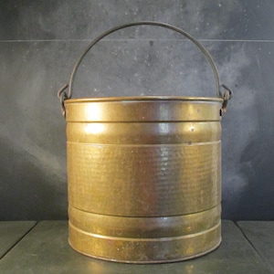 Puede incluir: Un cubo de cobre martillado con un asa de metal negro. El cubo mide aproximadamente 25 cm de alto y 20 cm de ancho.