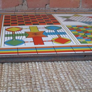 Op de afbeelding: Een vintage multi-game bord met een kleurrijk ontwerp. Het bord heeft een schaakbord, een slangen en ladders spel en een ludo-achtig spel. Het bord is gemaakt van metaal en heeft een versleten, rustieke uitstraling.