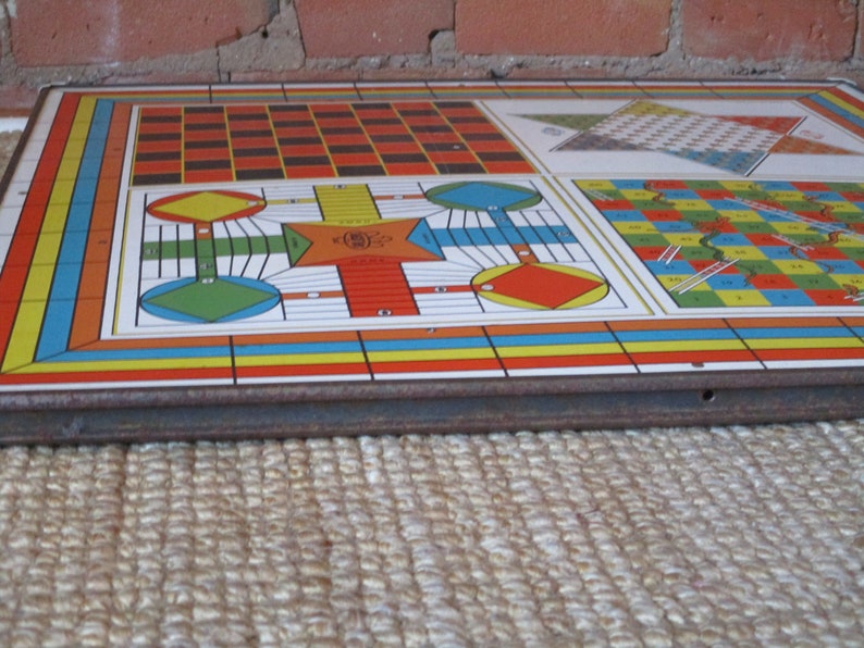 Op de afbeelding: Een vintage multi-game bord met een kleurrijk ontwerp. Het bord heeft een schaakbord, een slangen en ladders spel en een ludo-achtig spel. Het bord is gemaakt van metaal en heeft een versleten, rustieke uitstraling.