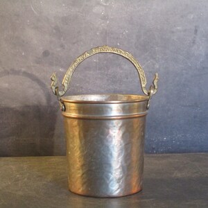 May include: A hammered copper bucket with a decorative brass handle. The bucket has a slightly distressed look and is perfect for a rustic or farmhouse style decor.
