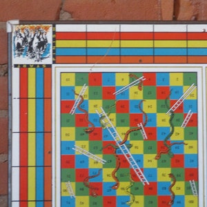 Op de afbeelding: Een vintage Slangen en Ladders bordspel met een kleurrijk raster van vierkanten, ladders en slangen. Het bord is gemaakt van metaal en heeft een rood, geel, blauw en groen kleurenschema. Het spel is in goede staat en heeft wat slijtage.