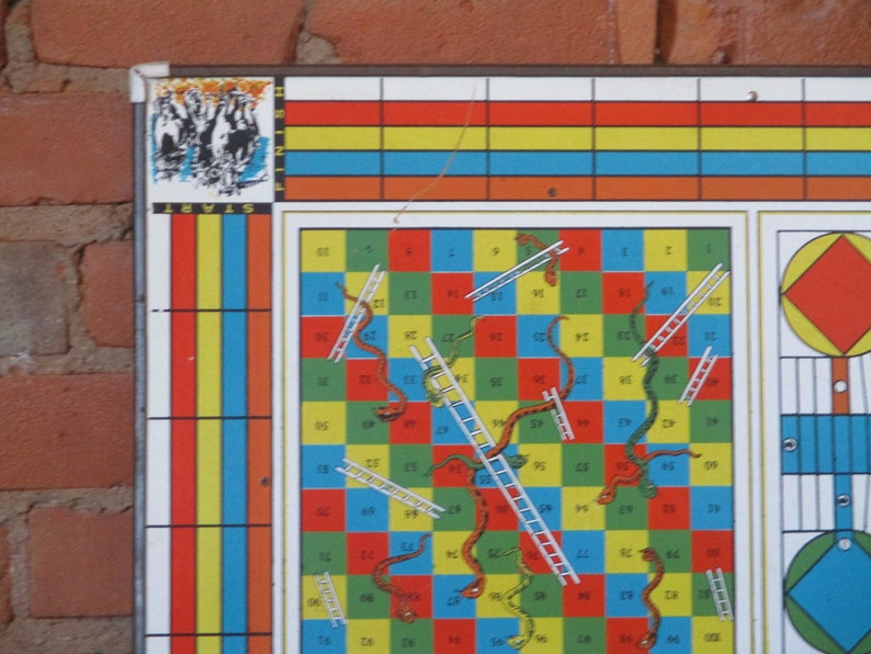 Op de afbeelding: Een vintage Slangen en Ladders bordspel met een kleurrijk raster van vierkanten, ladders en slangen. Het bord is gemaakt van metaal en heeft een rood, geel, blauw en groen kleurenschema. Het spel is in goede staat en heeft wat slijtage.