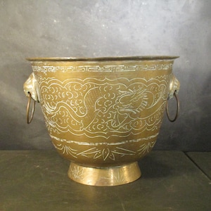 Puede incluir: Un macetero grande de metal dorado con dos asas de dragón. El macetero está decorado con un intrincado diseño de dragones y nubes.
