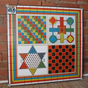 Op de afbeelding: Een vintage multi-game bord met vier verschillende spellen: Snakes and Ladders, Ludo, een stervormig spel en dammen. Het bord is gemaakt van metaal en heeft een kleurrijk ontwerp met rode, blauwe, gele, groene en zwarte vierkanten.