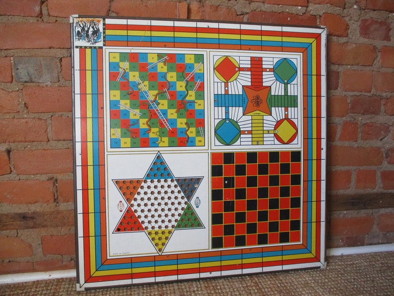 Op de afbeelding: Een vintage multi-game bord met vier verschillende spellen: Snakes and Ladders, Ludo, een stervormig spel en dammen. Het bord is gemaakt van metaal en heeft een kleurrijk ontwerp met rode, blauwe, gele, groene en zwarte vierkanten.