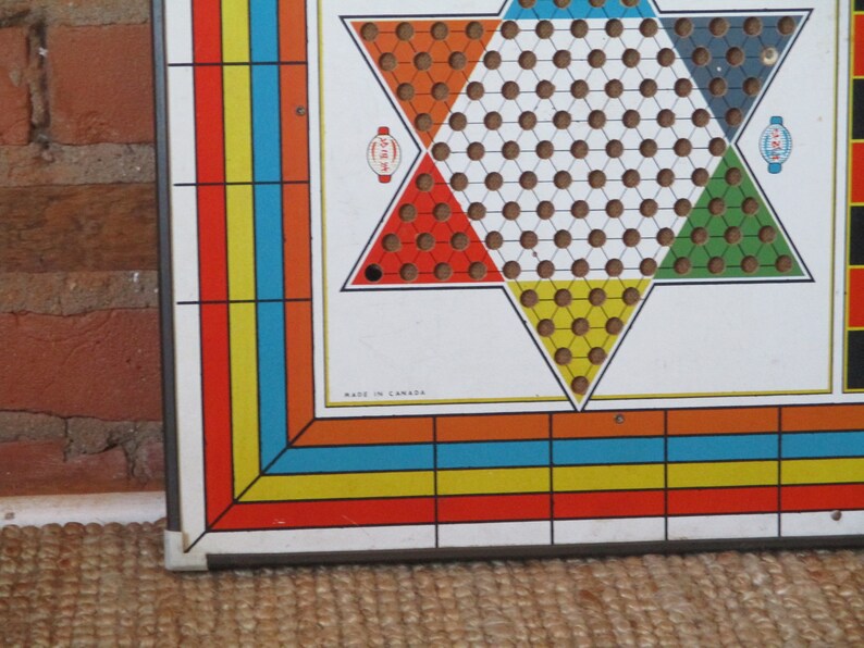 Op de afbeelding: Een vintage spelbord met een stervormig speelgebied. Het bord is gemaakt van metaal en heeft een witte achtergrond met rode, gele, blauwe en groene secties. Het bord is gelabeld "Made in Canada".