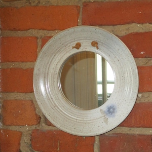 May include: A circular, off-white ceramic mirror with a speckled finish and a small blue floral accent. The mirror is mounted on a red brick wall. Two small, brown, decorative elements are attached to the top of the mirror.