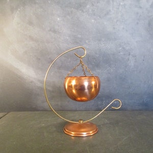 May include: A copper hanging planter with a curved stand. The planter is suspended from the stand by a chain.