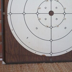 Op de afbeelding: Een vintage houten dartbord met een witte ronde target. De target is verdeeld in secties met de nummers 5, 10 en 15. Het midden van de target is een kleine zwarte cirkel.