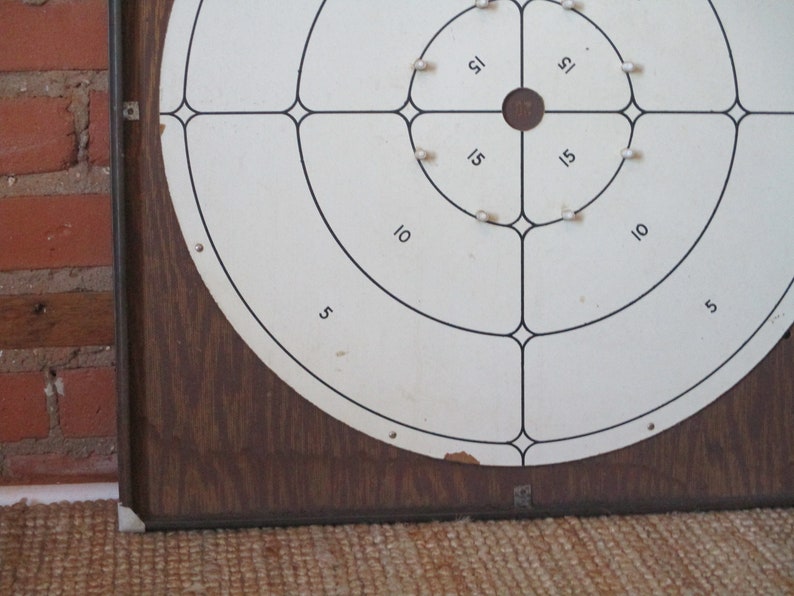 Op de afbeelding: Een vintage houten dartbord met een witte ronde target. De target is verdeeld in secties met de nummers 5, 10 en 15. Het midden van de target is een kleine zwarte cirkel.