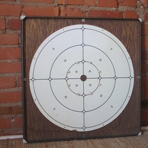 Op de afbeelding: Een vintage houten dartbord met een wit doel en zwarte ringen. Het doel heeft de nummers 5, 10, 15 en 21 erop gedrukt.