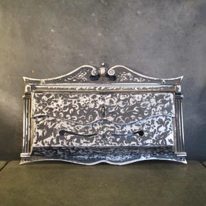 Può includere: Un mobile antico da parete in metallo con un motivo floreale decorativo. Il mobile ha una finitura argentata con accenti neri, un buco della serratura e due aperture curve. La parte superiore ha un design a scorrimento decorativo.