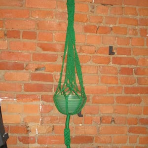 Peut inclure: Un porte-plante en macramé vert avec un pot en céramique vert suspendu à un crochet sur un mur en briques.