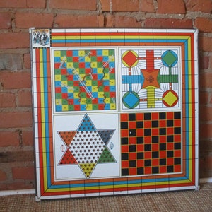 Op de afbeelding: Een vintage metalen spelbord met vier verschillende spellen: Snakes and Ladders, een dobbelspel, een stervormig spel en dammen. Het bord is geschilderd in felle kleuren, waaronder rood, geel, blauw, groen en zwart.