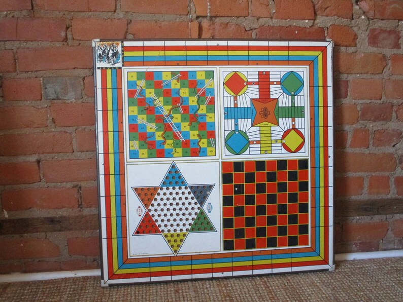 Op de afbeelding: Een vintage metalen spelbord met vier verschillende spellen: Snakes and Ladders, een dobbelspel, een stervormig spel en dammen. Het bord is geschilderd in felle kleuren, waaronder rood, geel, blauw, groen en zwart.