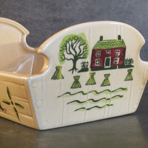 Może przedstawiać: Ceramiczny kosz na chleb z namalowanym na boku sceną wiejską. Scena przedstawia czerwony dom, zielone drzewo i stogi siana. Na przodzie kosza napisano "Chleb".