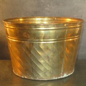 Puede incluir: Un macetero de metal dorado con un patrón decorativo retorcido. El macetero es redondo y tiene una parte superior ancha y abierta.