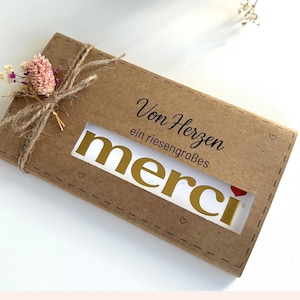 Pode incluir: Uma caixa de presente retangular de papel kraft com uma janela mostrando a palavra "merci" em letras douradas. A caixa é amarrada com barbante e decorada com flores secas. O texto "Von Herzen" está impresso na caixa.