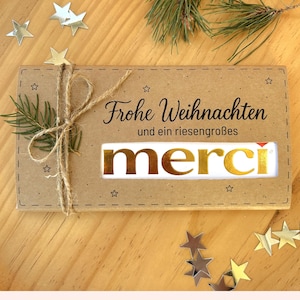 May include: A kraft paper gift box tied with twine, featuring the words "Frohe Weihnachten" and "merci" in gold lettering. The box is decorated with star-shaped confetti and a sprig of pine.