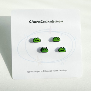 May include: A set of four green frog-shaped stud earrings on a white card. The earrings have black outlines and smiling faces. The card reads "CharmCharmStudio" and "Hypoallergenic Titanium Studs Earrings".