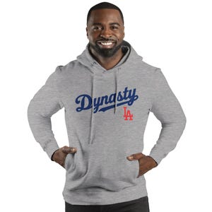 May include: A gray hooded sweatshirt with the word "Dynasty" in blue script and a red "LA" logo. The sweatshirt has a front pocket and a drawstring hood. The person is wearing the sweatshirt.