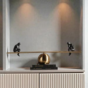 Puede incluir: Una estantería decorativa con dos esculturas negras de figuras sentadas sobre una barra dorada. Una esfera dorada se encuentra sobre una pila de libros, incluyendo "Les Misérables". La estantería está colocada contra una pared gris neutra.