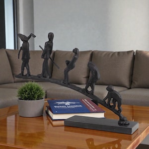 May include: A black metal sculpture depicting a series of stylized figures in various poses, arranged along a curved line. The sculpture is mounted on a rectangular black base and sits on a wooden table. A small potted plant and books are also on the table.