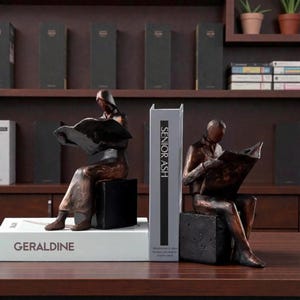 Könnte beinhalten: Bronzefarbene Buchstützen, die Figuren beim Lesen zeigen, sitzend auf schwarzen Würfelsockeln. Die Buchstützen stehen auf einer Holzoberfläche mit Büchern und Regalen im Hintergrund. Eine Buchstütze hat den Text "SENIOR ASH" auf dem Buchrücken.