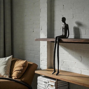 Op de afbeelding: Een zwarte metalen sculptuur van een zittende persoon met lange benen, geplaatst op een houten plank. De sculptuur staat tegen een witte bakstenen muur, met een bruine leren bank en kussens op de achtergrond.