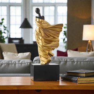 May include: A gold sculpture of a draped figure on a black rectangular base. The figure has a black head and shoulders, with flowing fabric. The sculpture is displayed on a wooden table in a living room setting.