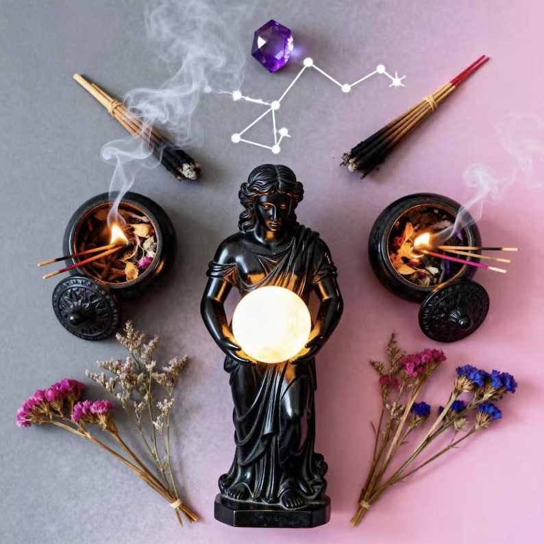 May include: A black statue of a woman holding a glowing orb, flanked by incense burners with burning incense sticks. Dried flowers and a purple crystal with a constellation outline are also present. The background is split between gray and pink.