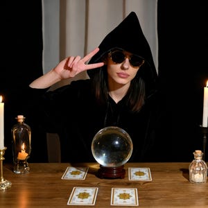 May include: A person wearing a black hooded cloak and sunglasses, gesturing with a peace sign. A crystal ball sits on a wooden table, surrounded by tarot cards and candles, creating a mystical atmosphere. A small glass bottle with a cork is also on the table.