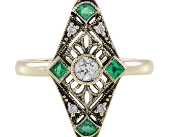 Può includere: Un anello dorato con un elemento centrale allungato a forma di diamante. L'anello presenta una pietra centrale rotonda e trasparente, circondata da pietre verdi quadrate e pietre trasparenti più piccole. Il design ha un motivo a filigrana aperta.