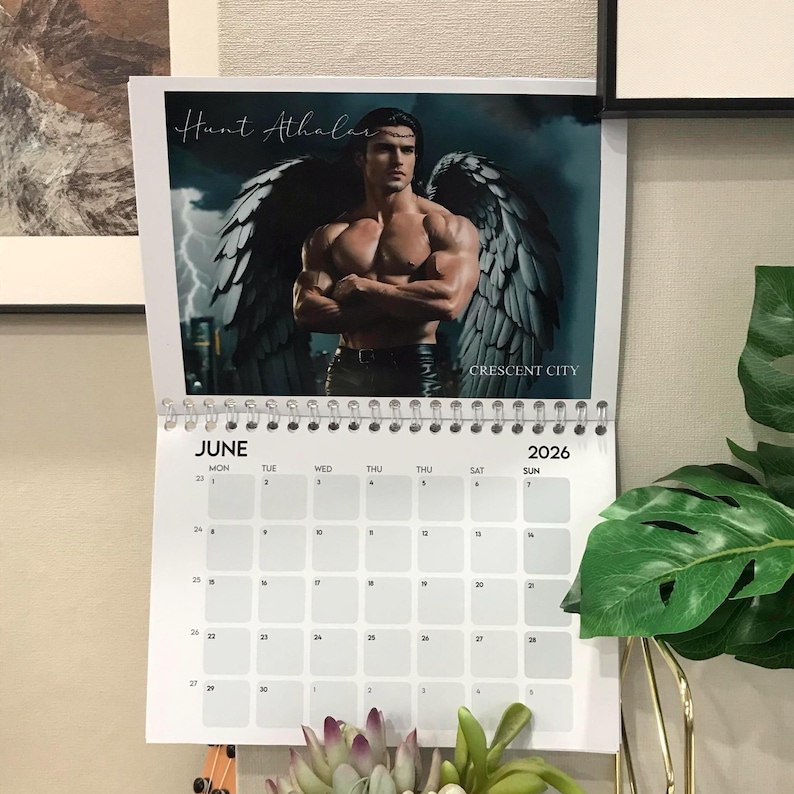 May include: A spiral-bound calendar for the year 2026, featuring a muscular man with angel wings. The calendar is open to the month of June, with a grid of dates and days. The title "Hunt Athalar" is visible.