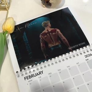May include: A spiral-bound calendar for 2026 featuring a shirtless man with a red wing tattoo on his back. The calendar shows the month of February with the text "FOURTH WING" and "Liam Maire". A yellow tulip and gold decor are in the background.